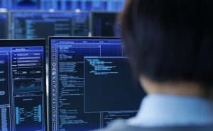 In the System Control Room Technical Operator Stands and Monitors Various Activities Showing on Multiple Displays with Graphics. Administrator Monitors Work of  Artificial Intelligence, Big Data Mining, Neural Network, Surveillance Project.